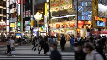 Belebte Kreuzung mit vielen Fußgängern, Neonreklamen und dicht stehenden Hochhäusern.