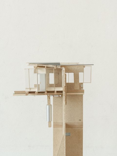 Eine detaillierte Seitenansicht des zweiten Modells, die die Konstruktion und die verwendeten Materialien fokussiert. Die Holzskelettkonstruktion, die verschiedenen Wandelemente aus Spanplatte, Holz und Glas sowie die Dachformen sind gut erkennbar. Der hohe Sockel aus Spanplatten bildet die Basis für das komplexe Modell. Das Bild ist vor dem gleichen neutralen Hintergrund aufgenommen wie die vorherigen Aufnahmen.