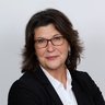 Das Bild zeigt ein professionelles Porträt von Dipl.-Ing. Kerstin Unger, Sachbearbeiterin in der Bauaufsicht der Stadt Bückeburg, vor einem hellen, neutralen Hintergrund. Sie steht leicht seitlich zur Kamera und hat die Arme vor dem Körper verschränkt. Sie trägt einen schwarzen Blazer über einer weißen Bluse. Ihr dunkles, schulterlanges Haar ist locker gestylt und leicht nach außen geföhnt. Sie trägt eine Brille mit dunklem Rahmen und dezente Ohrringe. Ihr Gesichtsausdruck ist ruhig und freundlich, mit einem leichten, geschlossenen Lächeln. Die Haltung wirkt selbstbewusst und sachlich, das Gesamtbild seriös und typisch für ein offizielles Verwaltungs- oder Presseporträt.