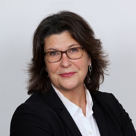 Das Bild zeigt ein professionelles Porträt von Dipl.-Ing. Kerstin Unger, Sachbearbeiterin in der Bauaufsicht der Stadt Bückeburg, vor einem hellen, neutralen Hintergrund. Sie steht leicht seitlich zur Kamera und hat die Arme vor dem Körper verschränkt. Sie trägt einen schwarzen Blazer über einer weißen Bluse. Ihr dunkles, schulterlanges Haar ist locker gestylt und leicht nach außen geföhnt. Sie trägt eine Brille mit dunklem Rahmen und dezente Ohrringe. Ihr Gesichtsausdruck ist ruhig und freundlich, mit einem leichten, geschlossenen Lächeln. Die Haltung wirkt selbstbewusst und sachlich, das Gesamtbild seriös und typisch für ein offizielles Verwaltungs- oder Presseporträt.