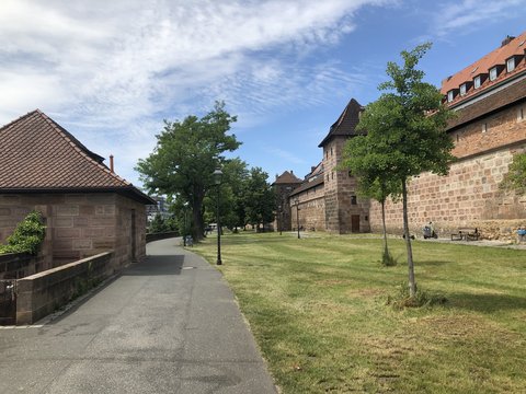 Historische Befestigungsanlage mit massiven Steinmauern und verschiedenen Gebäuden. Breite Grasfläche dazwischen. Schattige Bäume und Fußweg mit Laternen. Menschen spazieren. Blauer Himmel. Landschaftliche Grünfläche ohne intensive Bebauung.
