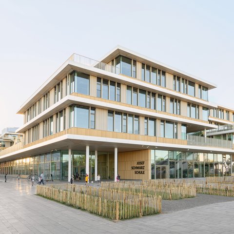„Außenansicht des mehrgeschossigen Holz- und Glasgebäudes der Josef Schwarz Schule mit terrassierten Ebenen und einem großen überdachten Eingangsbereich.“