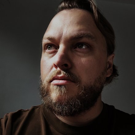 Porträt eines erwachsenen Mannes in Halbnahaufnahme, Blick nach rechts oben, kurzer Bart und Schnauze, kräftige Schattenzeichnung im Gesicht, dunkles T-Shirt, neutraler Hintergrund.