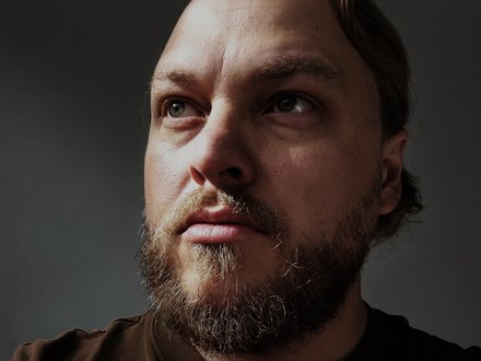 Porträt eines erwachsenen Mannes in Halbnahaufnahme, Blick nach rechts oben, kurzer Bart und Schnauze, kräftige Schattenzeichnung im Gesicht, dunkles T-Shirt, neutraler Hintergrund.