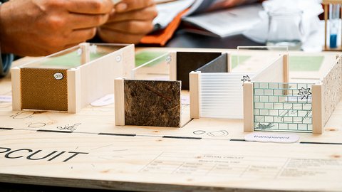 Nahaufnahme eines Architektur-Modellspiels auf einer Holzplatte. Kleine Wandelemente aus unterschiedlichen Materialien wie Holz, Wellpappe und Glas sind als Raumstrukturen aufgestellt.
