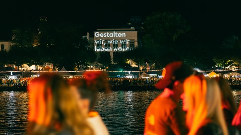 Mehrere Personen laufen nachts am Wasser entlang, im Hintergrund auf der anderen Flussseite ist ein Schild.