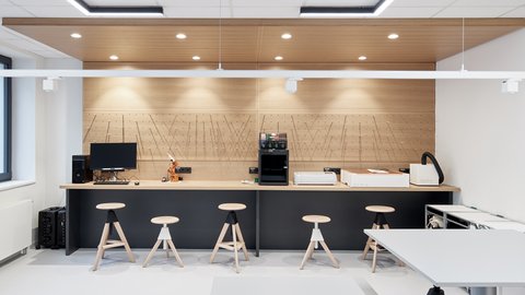 Moderner heller Raum mit langer Theke, vier Hockern, Computern und Kaffeemaschine, weiße Wände und Holzdecke mit Einbaustrahlern