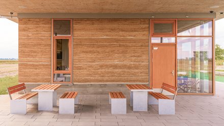 Überdachte Terrasse mit zwei Sitzgruppen vor Stampflehmwand, Glasfassade und Holztür, tagsüber.