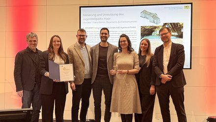 Sieben Personen auf einer Bühne bei einer Preisverleihung; eine hält eine Urkunde, eine weitere einen Glaspokal. Hinter ihnen eine Folie mit dem Titel „Sanierung und Umnutzung des Jugendstilparks Haar“, rotes Bühnenlicht.