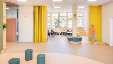 Heller, moderner Raum mit großen Fenstern, gelben Vorhängen, runden Sitzmöbeln und einer Person in orangefarbener Kleidung, die rechts vorbeigeht