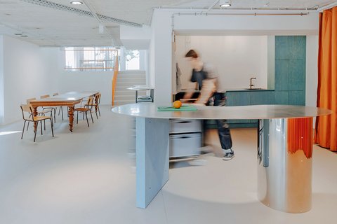 Heller, offener Innenraum mit geschwungener Metalltheke im Vordergrund, Küchenbereich dahinter und einem langen Holztisch mit Stühlen im Hintergrund.
