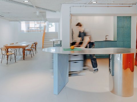Heller, offener Innenraum mit geschwungener Metalltheke im Vordergrund, Küchenbereich dahinter und einem langen Holztisch mit Stühlen im Hintergrund.