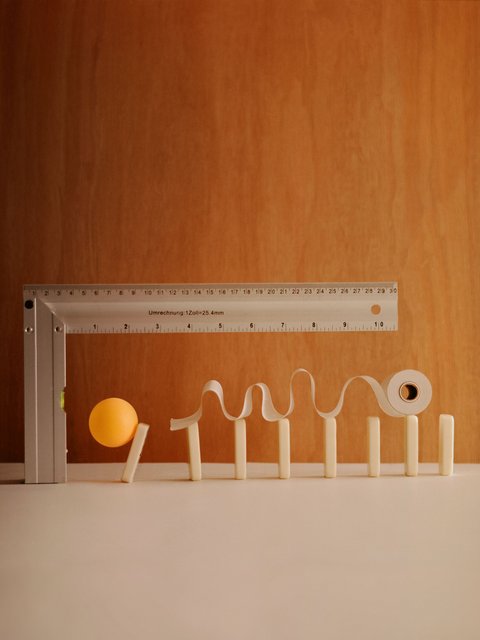 "Kreative Anordnung von beigen Holzelementen vor orangebraunem Hintergrund. Ein Messstab mit Zentimeterskala bildet die horizontale Achse. Weiße wellenförmige Formen und ein einzelner gelber Ball veranschaulichen Daten oder Messwerte graphisch."