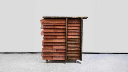 Mobiler Fassadenprototyp im Innenraum: Holzrahmen mit roten, überlappenden Schindeln in zwei Feldern, oben ein überstehendes Brett; auf Rollen.