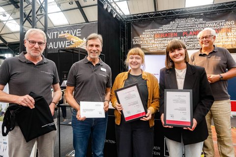 Mehrere Personen präsentieren gerahmte Auszeichnungen vor einem Informationsstand zur Architektur.