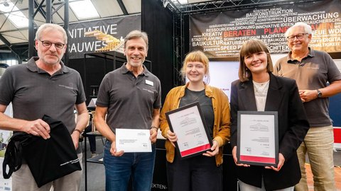 Mehrere Personen präsentieren gerahmte Auszeichnungen vor einem Informationsstand zur Architektur.