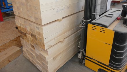 Werkhalle: Glatt gehobelte Holzträger zu einem Paket gebündelt; daneben die Gabel eines gelben Staplers.