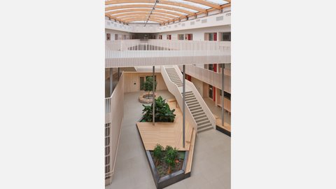 Innenansicht eines lichtdurchfluteten, modernen Atriums mit Holzgalerien, einer breiten Treppe und geometrisch angelegten Pflanzinseln.