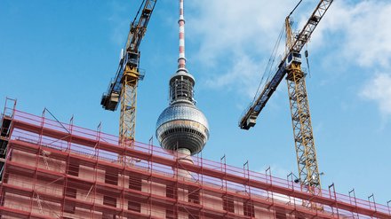 Baustelle in Berlin mit Gerüsten und zwei Baukränen im Vordergrund, dahinter der Fernsehturm vor blauem Himmel.