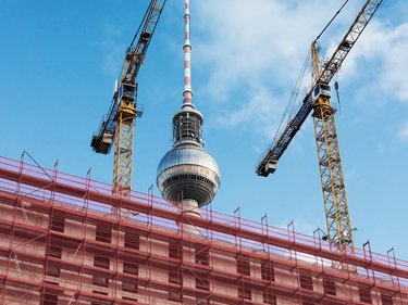 Baustelle in Berlin mit Gerüsten und zwei Baukränen im Vordergrund, dahinter der Fernsehturm vor blauem Himmel.