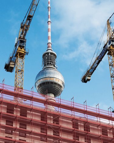 Baustelle in Berlin mit Gerüsten und zwei Baukränen im Vordergrund, dahinter der Fernsehturm vor blauem Himmel.