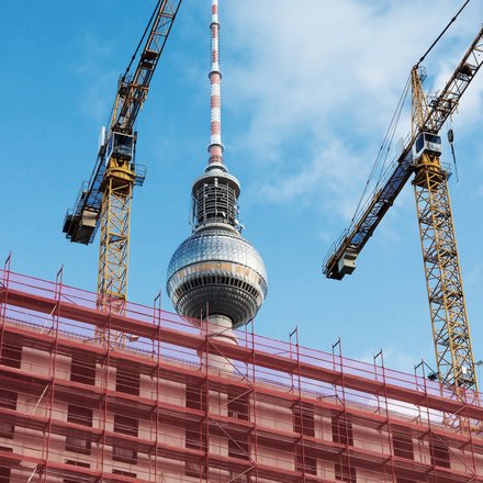 Baustelle in Berlin mit Gerüsten und zwei Baukränen im Vordergrund, dahinter der Fernsehturm vor blauem Himmel.