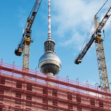 Baustelle in Berlin mit Gerüsten und zwei Baukränen im Vordergrund, dahinter der Fernsehturm vor blauem Himmel.