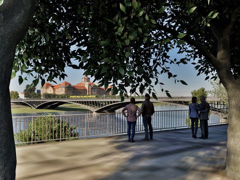 Eine Visualisierung zeigt einen begrünten Uferweg unter großen Bäumen mit Blick auf einen Fluss und eine moderne Brücke, mehrere Menschen bleiben stehen, lehnen am Geländer oder schauen gemeinsam auf das Wasser und die gegenüberliegende Stadtsilhouette, wodurch das Bild öffentlichen Raum am Wasser als Aufenthaltsort darstellt und für die Verbindung von Infrastruktur, Landschaft und sozialer Nutzung steht.