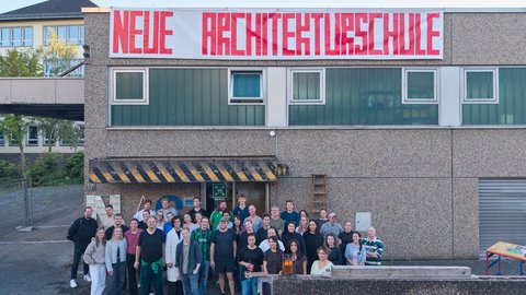 Gruppe von Menschen vor einem Gebäude mit einem großen Banner mit der Aufschrift 'NEUE ARCHITEKTURSCHULE'