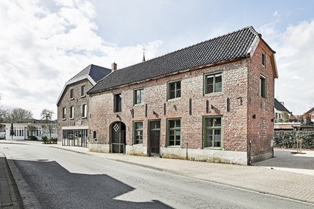 Außenansicht eines länglichen, roten Backsteinhauses mit grün gerahmten Fenstern und dunklem Ziegeldach an einer ruhigen Straße bei Tageslicht.