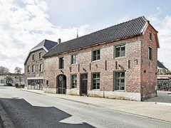 Außenansicht eines länglichen, roten Backsteinhauses mit grün gerahmten Fenstern und dunklem Ziegeldach an einer ruhigen Straße bei Tageslicht.