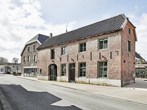 Außenansicht eines länglichen, roten Backsteinhauses mit grün gerahmten Fenstern und dunklem Ziegeldach an einer ruhigen Straße bei Tageslicht.