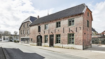 Außenansicht eines länglichen, roten Backsteinhauses mit grün gerahmten Fenstern und dunklem Ziegeldach an einer ruhigen Straße bei Tageslicht.
