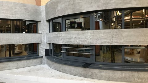 Rundes Gebäude in Grau mit zwei Reihen an Fenstern in denen Spiegelungen zu erkennen sind 