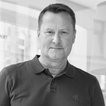 Schwarz-Weiß-Porträt einer Person in dunklem Poloshirt vor einer Glaswand in einem hellen Bürogebäude.