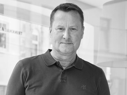 Schwarz-Weiß-Porträt einer Person in dunklem Poloshirt vor einer Glaswand in einem hellen Bürogebäude.