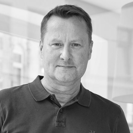 Schwarz-Weiß-Porträt einer Person in dunklem Poloshirt vor einer Glaswand in einem hellen Bürogebäude.