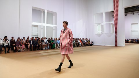 "Ein Model läuft über einen hellen Laufsteg in einem minimalistisch gestalteten Raum mit weißen Wänden und geometrischen Wandöffnungen. Es trägt ein rot-weiß kariertes Kleid. Das Publikum sitzt entlang der Seiten auf einfachen Bänken."