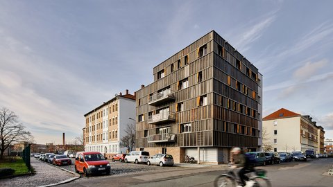 „Modernes Wohngebäude mit markanter dunkler Fassade und orangen Fensterläden an einer Straßenecke in einem städtischen Wohnviertel.“