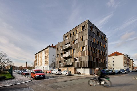 „Modernes Wohngebäude mit markanter dunkler Fassade und orangen Fensterläden an einer Straßenecke in einem städtischen Wohnviertel.“