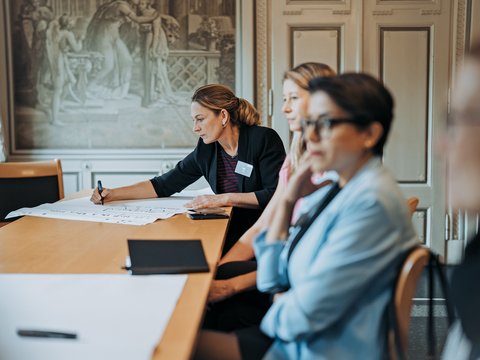 "Eine Frau im schwarzen Blazer mit Namensschild schreibt auf weißem Papier an einem Holztisch. Sie sitzt vor zwei anderen Frauen, die zuhören. Im Hintergrund klassisches Klassenzimmer mit dekorativem Wandgemälde. Schulungssituation oder Moderationsrunde."