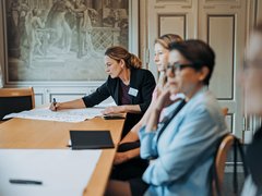 "Eine Frau im schwarzen Blazer mit Namensschild schreibt auf weißem Papier an einem Holztisch. Sie sitzt vor zwei anderen Frauen, die zuhören. Im Hintergrund klassisches Klassenzimmer mit dekorativem Wandgemälde. Schulungssituation oder Moderationsrunde."