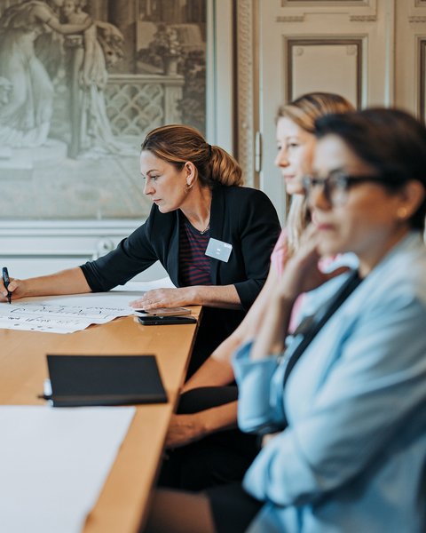 "Eine Frau im schwarzen Blazer mit Namensschild schreibt auf weißem Papier an einem Holztisch. Sie sitzt vor zwei anderen Frauen, die zuhören. Im Hintergrund klassisches Klassenzimmer mit dekorativem Wandgemälde. Schulungssituation oder Moderationsrunde."