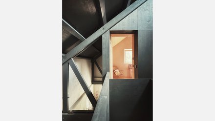 Blick unter das Dach: Dunkles Stahltragwerk mit Diagonalen, Durchblick in einen warm roséfarbenen Raum mit Sessel und Fenster.