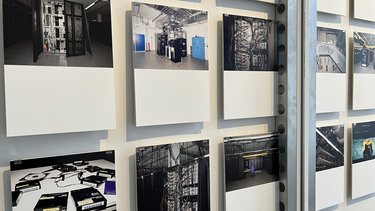 Mehrere Fotografien hängen einzeln mit Abstand an einer Wand. Zu sehen sind historische und moderne Technikräume: Rechenzentren, Schaltschränke, Kabelbündel und alte Computersysteme. Die Bilder wirken kühl, dunkel und komplex. Die Installation macht die sonst verborgene Infrastruktur des Internets sichtbar und erfahrbar.