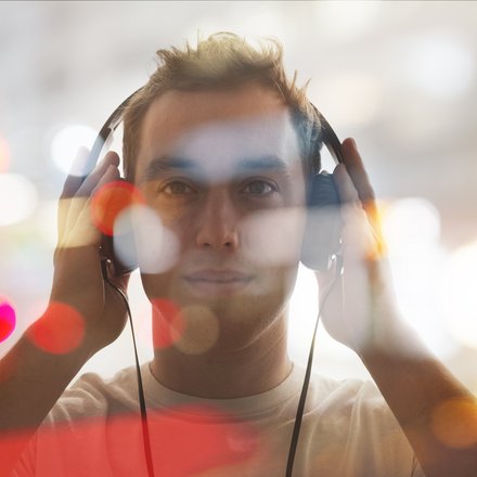 Porträt eines jungen Mannes, frontal, mit großen Kopfhörern, beide Hände an den Hörmuscheln; das Bild ist mit unscharfen Bokeh‑Lichtpunkten überlagert, warme Farbtupfer und heller, verschwommener Hintergrund.