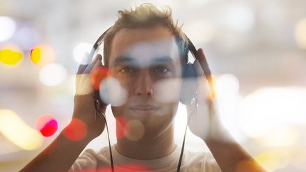 Porträt eines jungen Mannes, frontal, mit großen Kopfhörern, beide Hände an den Hörmuscheln; das Bild ist mit unscharfen Bokeh‑Lichtpunkten überlagert, warme Farbtupfer und heller, verschwommener Hintergrund.
