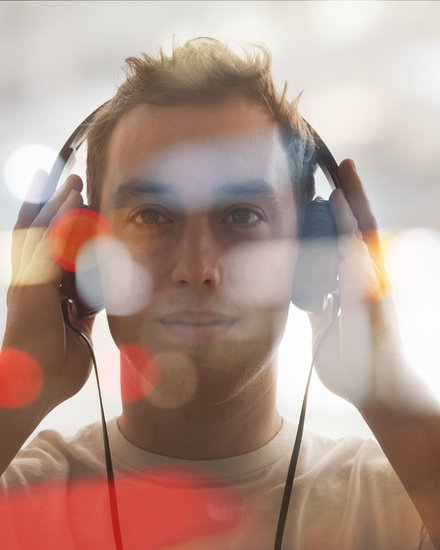 Porträt eines jungen Mannes, frontal, mit großen Kopfhörern, beide Hände an den Hörmuscheln; das Bild ist mit unscharfen Bokeh‑Lichtpunkten überlagert, warme Farbtupfer und heller, verschwommener Hintergrund.