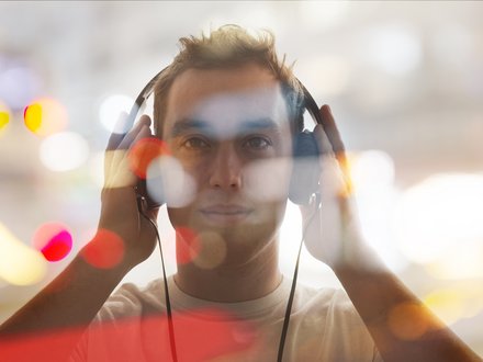 Porträt eines jungen Mannes, frontal, mit großen Kopfhörern, beide Hände an den Hörmuscheln; das Bild ist mit unscharfen Bokeh‑Lichtpunkten überlagert, warme Farbtupfer und heller, verschwommener Hintergrund.