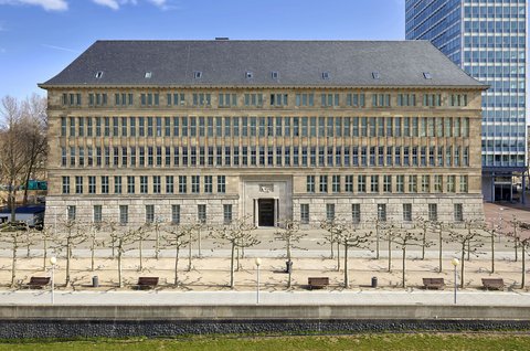 Fassade eines mehrstöckigen historischen Gebäudes mit symmetrischen Fenstern und einem zentralen Eingang, davor ein gepflasterter Platz mit kahlen Bäumen